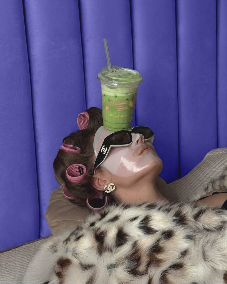 A woman in a sheet mask and rollers balancing a matcha